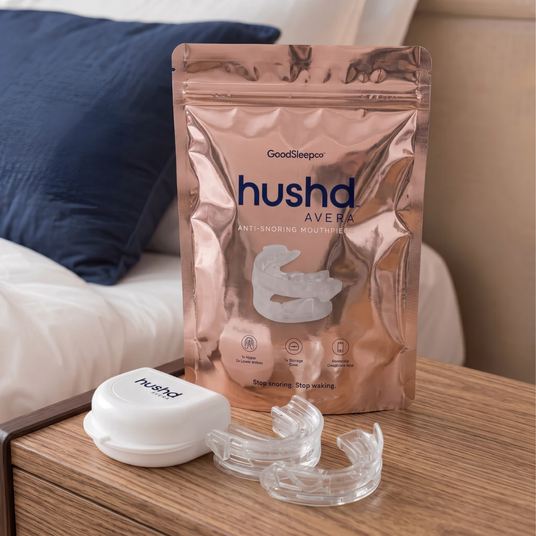 A metallic rose-gold pouch of hushd Avera anti-snoring mouthguards stands on a wooden bedside table next to a white carrying case and two clear mouthguards, with a bed and blue pillow in the background.