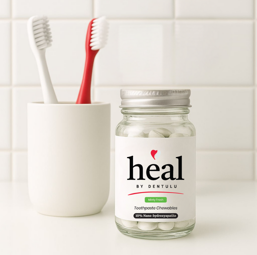 Jar of 'heal by Dentulu' toothpaste chewables with a toothbrush in a holder on a tiled bathroom counter.